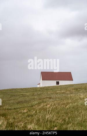 Ein einsames weißes Häuschen mit einem minimalistischen roten Dach, typisch für skandinavische Länder, auf einem grasbewachsenen Hügel. Wolkiger und grauer Tag. Einsamkeit Stockfoto