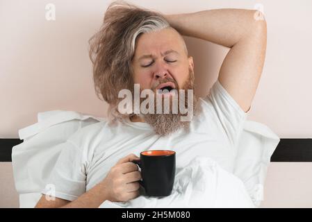 Gähnender bärtiger Mann, der morgens Kaffee trinkt und im Bett liegt. Ein Typ, der aufwachen will. Nahaufnahme Stockfoto