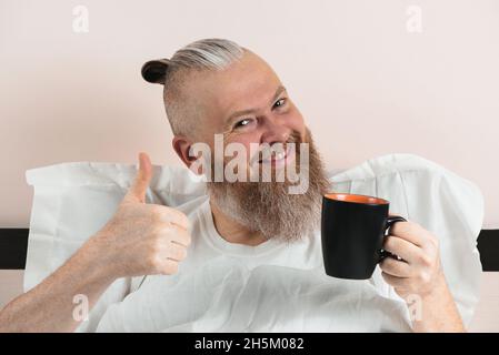 Glücklicher bärtiger Mann, der morgens Kaffee im Bett genoss und dabei den Daumen nach oben zeigte. Lächelnder Hipster-Typ beginnt den Tag. Nahaufnahme Stockfoto