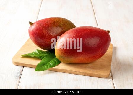 Rote reife Palmer Mangos auf einem Schneidebrett über weißem Holztisch. Bereit, süße leckere Mango zu essen. Köstliche tropische Früchte der Mangifera Indica. Stockfoto