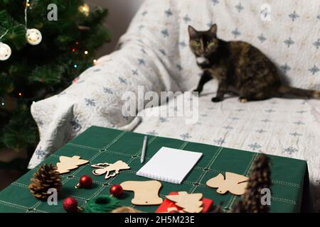 Ein Notizbuch mit einem Stift liegt auf dem Tisch mit einer grünen Tischdecke, die Katze sitzt in festlicher Atmosphäre auf der Couch. Selektiver Fokus. Vorbereitung für den Stockfoto