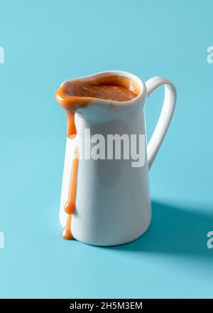 Nahaufnahme mit einem weißen Krug mit Karamellsauce auf einem blauen Tisch. Köstliche hausgemachte Karamellsauce tropft auf einem weißen Porzellan-Creamer. Stockfoto