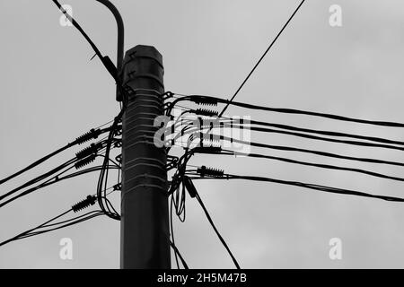 Spitze eines Mastes mit Stromkabeln unter bewölktem Himmel, schwarz-weißes Industriefoto Stockfoto