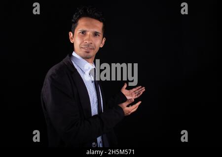 Geschäftsmann zeigt, erklärt oder präsentiert etwas mit seinen beiden Armen. Junger Geschäftsmann zeigt mit seinen Händen, isoliert auf schwarzem Hintergrund. Stockfoto