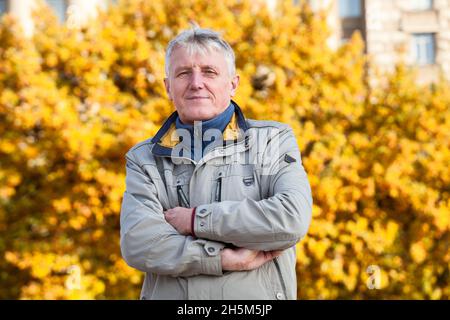 Älterer lächelnder kaukasischer Mann mit gefalteten Armen auf der Brust, gelbes Baumblatt als Hintergrund, Herbstsaison Stockfoto