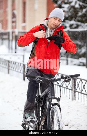 Der Lieferer steckt sein Smartphone in seine Jackentasche, während er in der Wintersaison auf dem Fahrrad sitzt Stockfoto