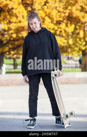 Ganzkörperportrait einer jungen Frau mit Longboard in den Händen, schwarzem Hoodie und trendigen Jeans, Herbstsaison mit gelben Blättern Stockfoto