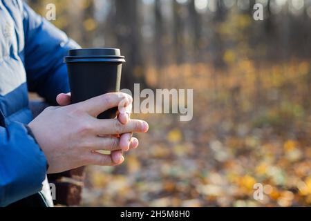 Im Herbst hält ein Mann einen Einweg-Papierbecher mit einem Getränk im Park. Das Konzept der Getränke, Tee, Kaffee, Herbst, Aufwärmen, Ruhe. Stockfoto