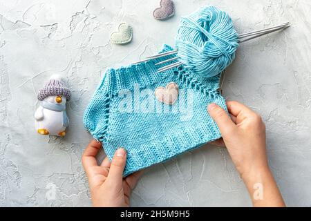 Kinderhände mit einem gestrickten T-Shirt für ein Spielzeug aus türkisfarbenem Baumwollgarn. Eine Kugel aus Garn und Stricknadeln. Hobbys. Die Entwicklung von Stockfoto