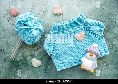 Das Konzept des Strickens von Kleidung für Spielzeug und Babys. Ein gestricktes T-Shirt aus türkisfarbenem Baumwollgarn. Eine Kugel aus Garn, Stricknadeln, Spielzeug und Samt Stockfoto