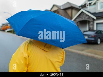 Frau mit blauem Regenschirm, die im Regen auf einer Straße läuft. Nicht erkennbare Person in gelber Regenjacke mit Regenschirm. Stockfoto