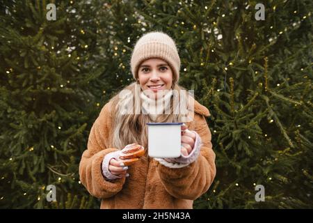 Cute Girl Holding Tea Cup und Sweet Bun lächeln im Winterwald. Verschneite Wintertage. Schönes Mädchen. Mädchen mit heißem Getränk in den Händen Stockfoto