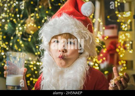 Kleine lustige Weihnachtsmann Kind essen Weihnachtskekse und ein Glas Milch auf Weihnachtsbaum Hintergrund. Stockfoto