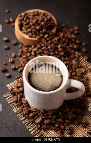 Kaffeebohnen auf einem rustikalen schwarzen Tisch verteilt und weiße Lungo-Tasse mit heißem Kaffeegetränk. Nahaufnahme des Bildes Stockfoto