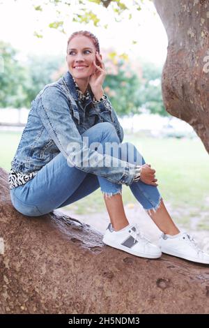 Junge, fröhliche, kaukasische Rotschopf-Frau, die in der Natur auf einem Ast sitzt, lächelt verjüngt, schaut weg Stockfoto