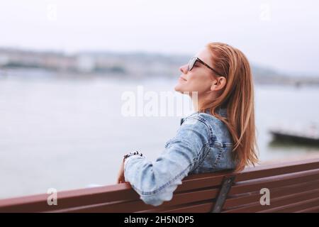 Junge ausgeglichene Rothaarige kaukasische Frau, die auf einer Bank am Flussufer sitzt und meditiert Stockfoto