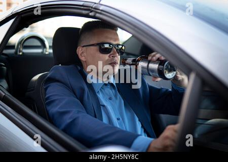 Ein Mann in einem teuren Anzug trinkt am Steuer eines Autos Bier, was die Gefahr eines Notfalls verursacht. Ein Geschäftsmann trinkt während der Fahrt. Betrunkener Fahrer Konz Stockfoto