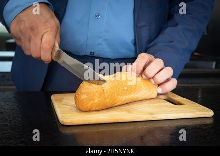 Die Hände des Mannes schneiden Brot auf dem Holzbrett. Ein einziger Geschäftsmann macht sich ein Sandwich in der Küche Stockfoto