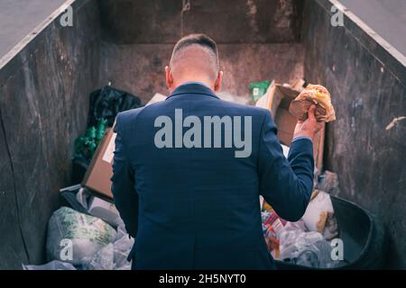 Ein verarmter Geschäftsmann rumpelt in einem Mülleimer auf der Suche nach Nahrung. Ein obdachloser Obdachloser in einem Anzug hält im Stehen einen Hamburger in der Hand Stockfoto