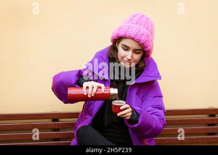 Eine positive Frau verbringt bei frostigen Wetterbedingungen Zeit im Freien, trinkt ein heißes Getränk aus einer Thermoskanne, lächelt, fröhlich in Oberbekleidung gekleidet. Wintersport und Reisekonzept Stockfoto