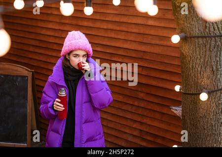 Eine positive Frau verbringt bei frostigen Wetterbedingungen Zeit im Freien, trinkt ein heißes Getränk aus einer Thermoskanne, lächelt, fröhlich in Oberbekleidung gekleidet. Wintersport und Reisekonzept Stockfoto