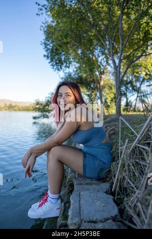 Junge asiatische Frau im strahlenden Sonnenlicht, die am Ufer eines idyllischen Sees sitzt Stockfoto