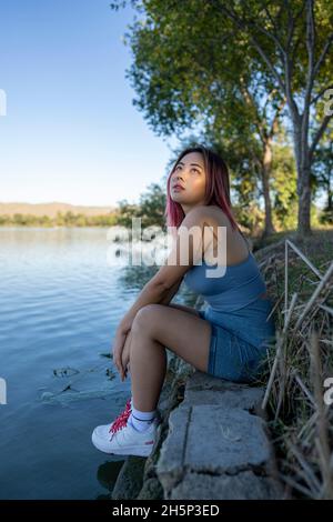 Junge asiatische Frau im strahlenden Sonnenlicht, die am Ufer eines idyllischen Sees sitzt Stockfoto