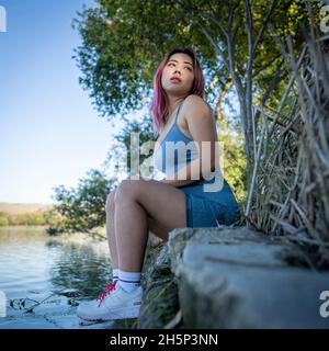 Junge asiatische Frau im strahlenden Sonnenlicht, die am Ufer eines idyllischen Sees sitzt Stockfoto