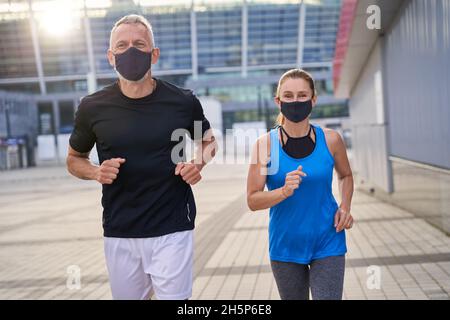 Sportives Paar mittleren Alters joggt morgens mit Schutzmasken. Covid19 Verantwortungsvolles Verhalten Stockfoto