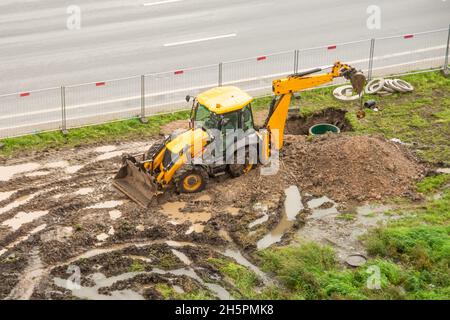 Notfallreparaturen der städtischen Hauptrohre auf dem nahegelegenen Rasen. Der arbeitende Traktorbagger gräbt einen Grubengraben vor Ort eines Heizungsausfalls Stockfoto