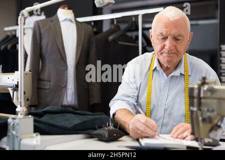 Älterer Schneider verwendet Notizblock, um in der Werkstatt Messungen der Jacke aufzuzeichnen Stockfoto