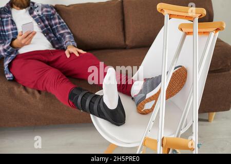 Junger Mann mit gebrochenem Bein oder verletztem Fuß sitzt auf der Couch und benutzt Handy Stockfoto