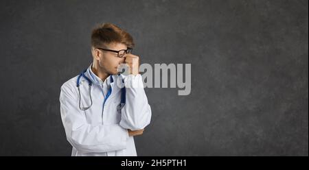 Porträt eines jungen erschöpften männlichen Arztes, der auf dem Hintergrund einer grauen Betonwand steht. Stockfoto