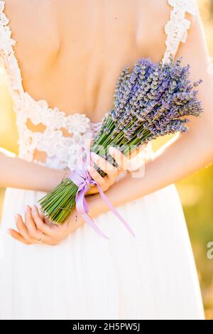 Braut in einem weißen Spitzenkleid steht auf einem Feld und hält einen Lavendelstrauß, der mit einem Fliederband hinter ihrem Rücken gebunden ist. Nahaufnahme Stockfoto