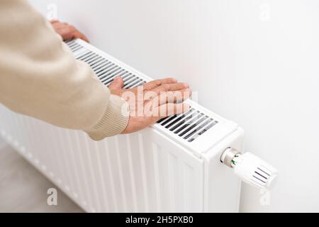 Ein älterer Mann erstarrte in seinen Händen, ein Mann erwärmt seine Hände in der Nähe eines Heizkörpers in einer Wohnung, ein Mann erstarrt in einer Wohnung. Stockfoto