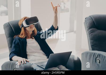 Weiblich im VR-Headset, zeigt mit dem Finger und berührt 3d-Objekte, während sie aus der Ferne auf dem Laptop arbeiten Stockfoto