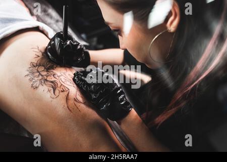 Professionelle weibliche Tattoo-Künstlerin macht ein Tattoo im Studio, Nahaufnahme. Dunkle Fotografie Stockfoto