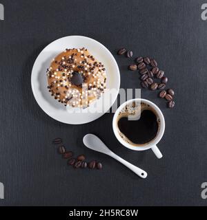 Donut dekoriert mit weißen und dunklen Schokoladenkugeln in Teller und Tasse Kaffee auf schwarzem Stein Hintergrund, Draufsicht Stockfoto