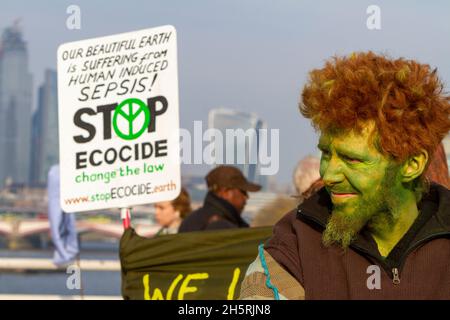 Straßenfoto des Protesters zum Klimawandel mit grün gemaltem Gesicht rechtes Bild. Großes Plakat links mit Londoner Gebäuden im Hintergrund. Stockfoto