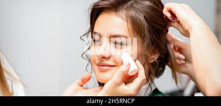 Makeup-Künstler, der in einem Schönheitssalon ein cremefarbenes Grundrohr auf die Wange der jungen kaukasischen Frau aufführt Stockfoto