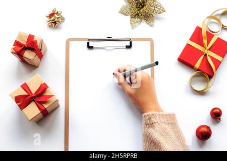 Frauenhand mit Bleistift in Notizbuch auf weißem Tisch mit Weihnachtsgeschenken und -Dekorationen. Draufsicht, flach liegend. Modell Stockfoto