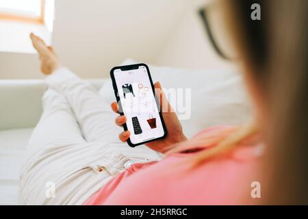 Junge Frau mit Online-Shop-App kaufen Kleidung. Frau zu Hause, die im Wohnzimmer mit dem Smartphone auf dem Bett liegt, kauft im Internet-Shop Kleidung. Stockfoto