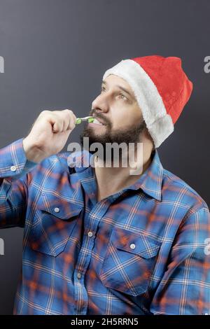 Bärtiger Mann mit Weihnachtsmütze und kariertem Hemd, der die Zähne putzt und nach oben schaut Stockfoto