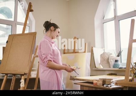 Nette Frau malt in einer Kunstwerkstatt auf Leinwand. Künstler, der ein Bild erstellt. Kunstschule oder Atelier. Arbeiten Sie mit Farben, Pinsel und Staffelei. Hobby und Freizeit Stockfoto
