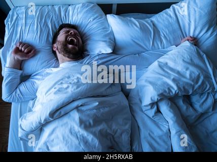 Der junge bärtige Mann im weißen Schlafanzug wacht aus dem Schlaf auf, er gähnt und streckt sich, während er im Bett liegt. Stockfoto