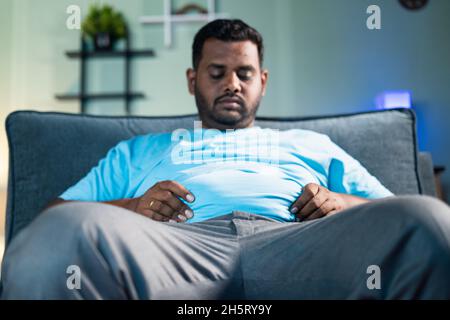 Trauriger Obase-Mann für schwere Bauchfett enttäuscht, indem sie die Hände beim Sitzen auf dem Sofa zu Hause - Konzept von Übergewicht, Krankheit, leiden an Überschuss Stockfoto