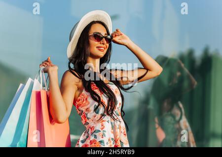 Stilvolle, glückliche junge Frau in Kleid und Sonnenbrille, die während des Einkaufsprozesses am Black Friday und des Konsumerismus mit farbenfrohen Einkaufstaschen in der Nähe des Stores spazieren geht Stockfoto