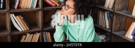Nachdenkliche afroamerikanische Studentin in Brillen, die in der Bibliothek wegschaut, Banner Stockfoto