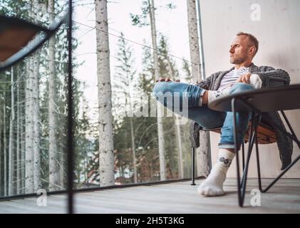Porträt eines Mannes mittleren Alters, der offene Strickjacke, Jeans und warme Socken trägt und auf dem Balkon des Waldhauses sitzt und die frische Luft genießt. Alltagslebenszyll Stockfoto