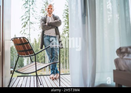 Ganzkörperportrait eines aufrichtig lächelnden Mannes mittleren Alters, gekleidet in einer offenen Strickjacke, Jeans auf dem Balkon des Waldhauses und die frische Luft genießen. Immer Stockfoto